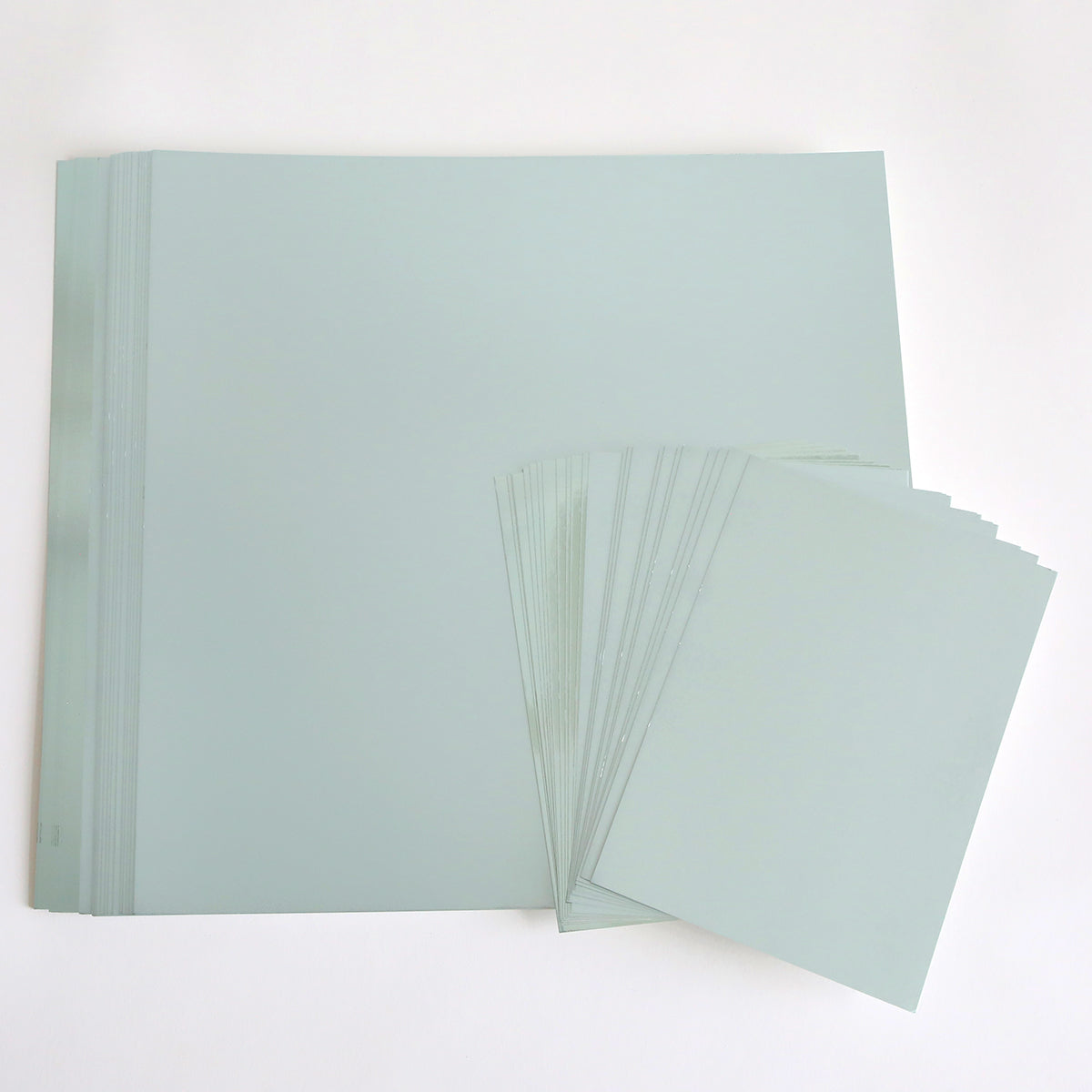 A stack of large, ice blue foil cardstock sheets lies next to a smaller stack of similar sheets, all arranged on a white surface—ideal for papercraft cardstock projects where both shiny and matte finishes from the Ice Blue Foil Cardstock Bundle are desired.
