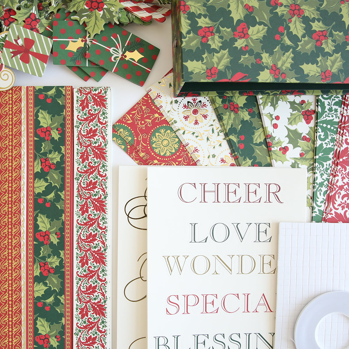 A collection of festive wrapping paper and gift boxes with holly and decorative patterns. Adjacent are sheets of white paper featuring words like "CHEER," "LOVE," "WONDER," and "SPECIAL" from the Simply Noel Scrapbooking Kit, perfect for adding a touch of holiday spirit to your scrapbooking projects.