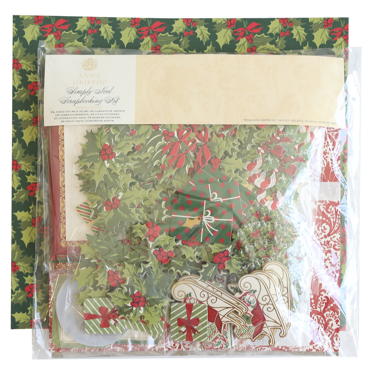 A packaged Simply Noel Scrapbooking Kit lies on festive holly-patterned paper, containing decorative elements like wreaths, bells, and stockings.