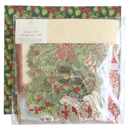 A packaged Simply Noel Scrapbooking Kit lies on festive holly-patterned paper, containing decorative elements like wreaths, bells, and stockings.