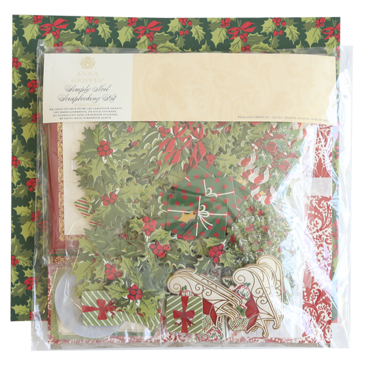 A packaged Simply Noel Scrapbooking Kit lies on festive holly-patterned paper, containing decorative elements like wreaths, bells, and stockings.