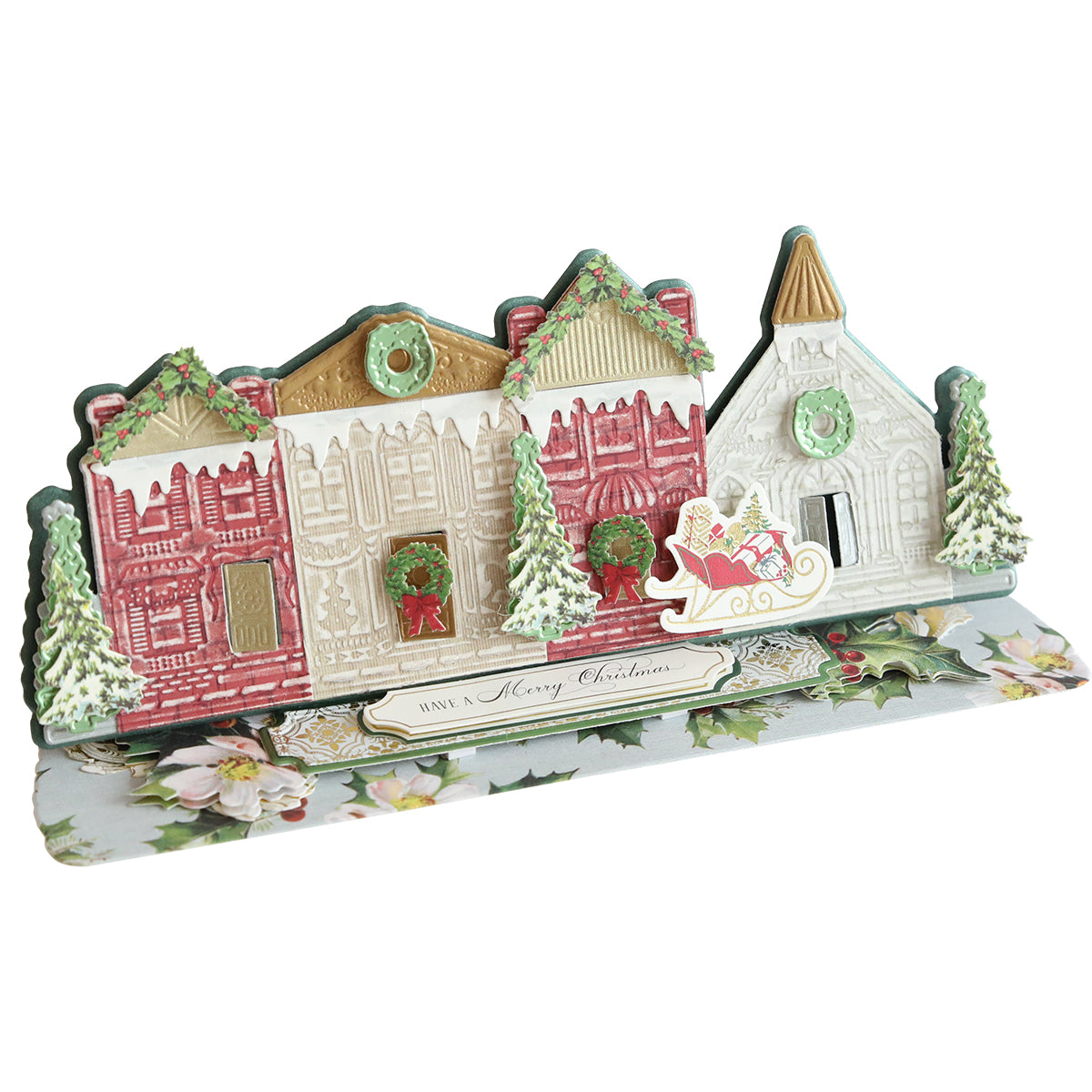 A decorative Christmas card featuring a 3D pop-up of a festive village scene with buildings, Christmas trees, and a sleigh. The card reads "Have a Merry Christmas" at the bottom and is perfect for anyone who loves Christmas crafts and the intricate details of the Village Easel Dies.