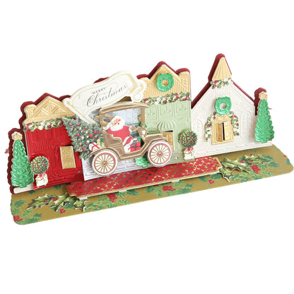 Village Easel Dies depicting Santa in a car with gifts, surrounded by a Christmas village of festive houses, wreaths, and decorated trees, set on a gold base with holly designs. Perfect for those who enjoy Christmas crafts.