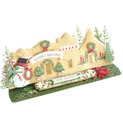 A festive pop-up card depicting a holiday village with a snowman, trees, wreaths, and a "Season's Greetings" banner. The card also contains a folded tag that says "Happy Holidays," resembling intricate Christmas crafts using the Village Easel Dies.