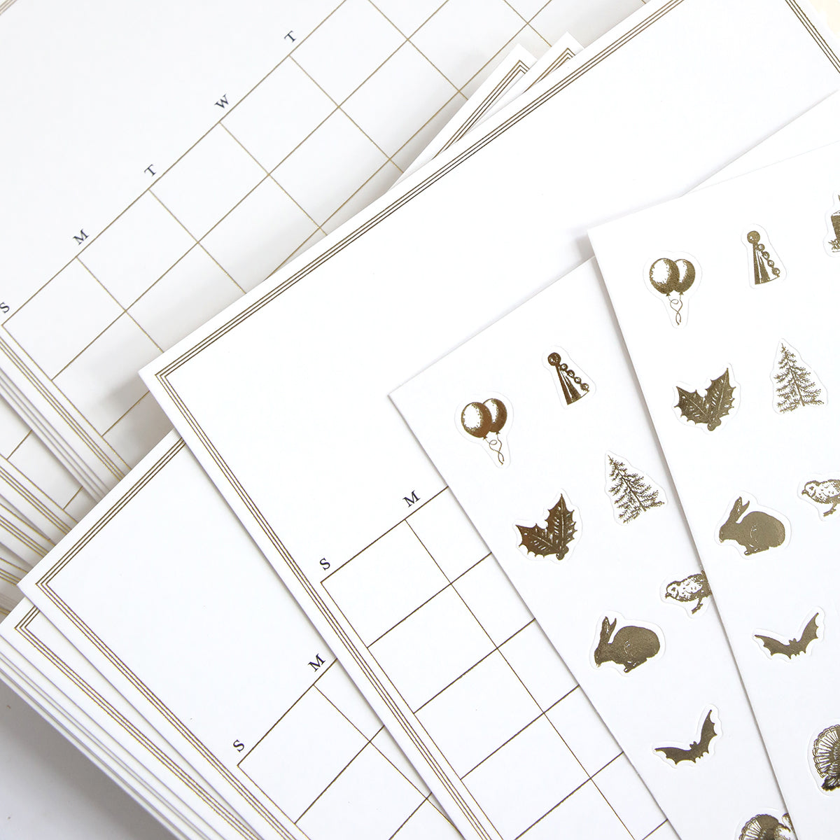 A collection of calendar sheets, accompanied by gold sticker sheets showcasing designs like balloons, trees, and animal shapes, from the Creative Calendars Refill Kit.