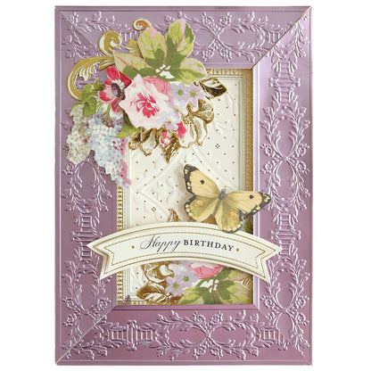 The 12" Cut and Emboss Folder Autoship offers an ornate lavender birthday card adorned with embossed floral designs reminiscent of scrapbook pages, featuring a yellow butterfly and "Happy Birthday" text on a banner.