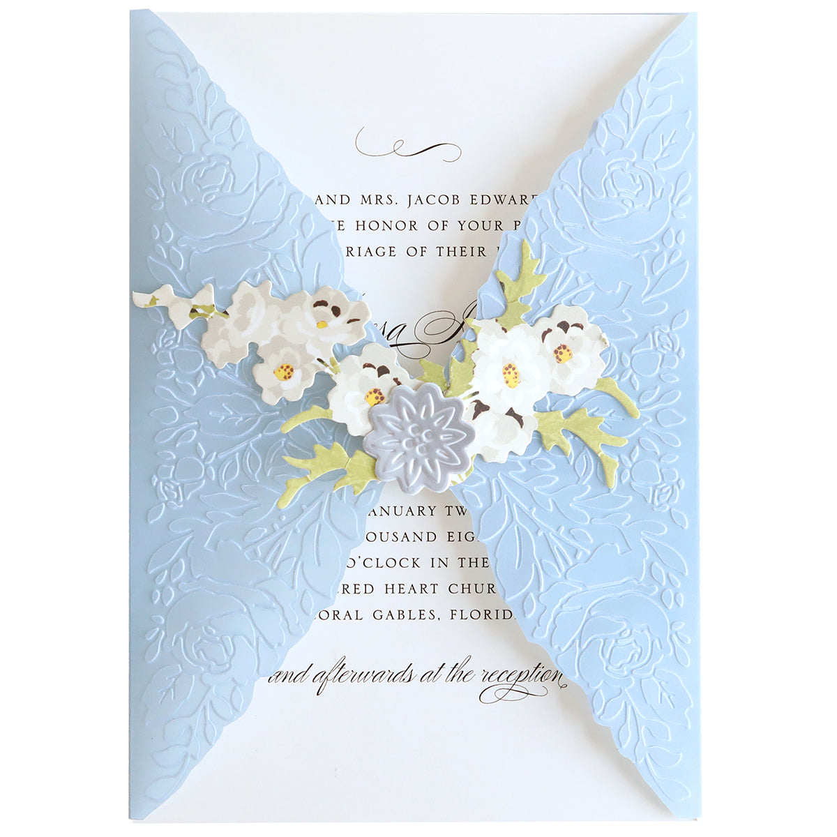 A wedding invitation crafted with the Envelope Seal Cut and Emboss Folders, featuring a blue floral embossed cover. It is partially open to reveal printed text inside and is adorned with delicate white and yellow flowers in the center, making this exquisite piece a testament to handmade creations.