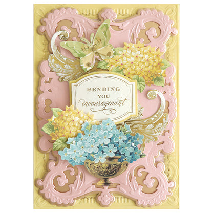 A handmade card featuring a decorative frame die from the Album Frame Dies collection, embellished with embossed pink patterns, yellow hydrangeas, a butterfly, and a bowl of blue flowers. This design beautifully conveys the heartfelt message: "Sending You Encouragement.