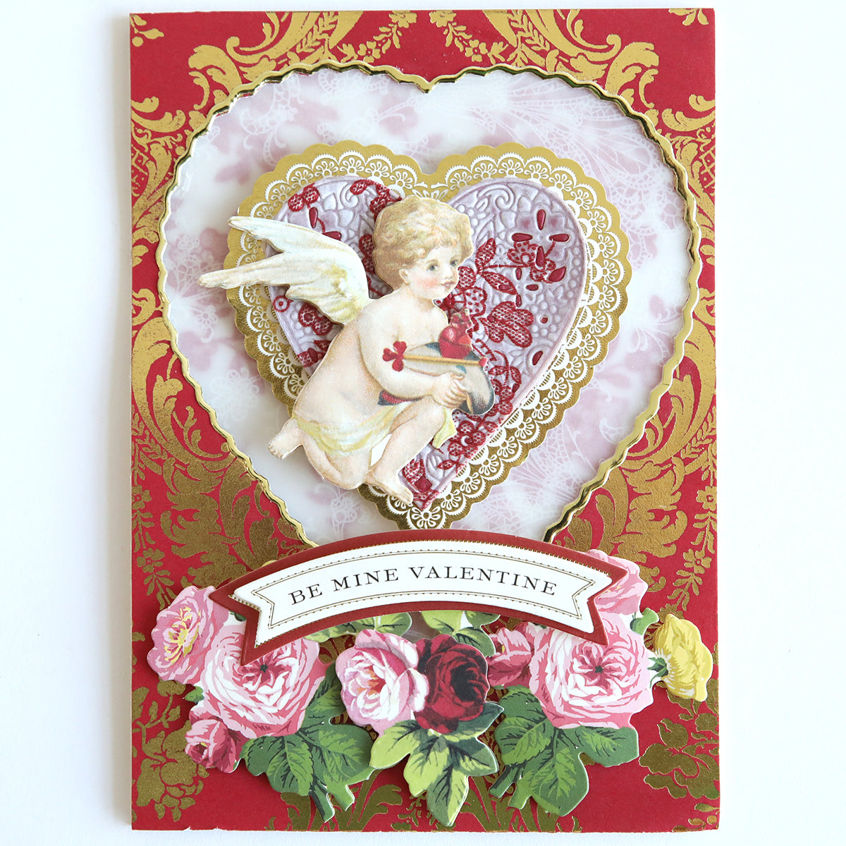 Valentine's card designed with an intricate heart pattern and a cherub holding a red heart, embellished with roses at the bottom. It features a 12" x 12" vellum sheet from the Red and White Lace Vellum Papers collection, adding an elegant transparent effect, with the caption "Be Mine Valentine.