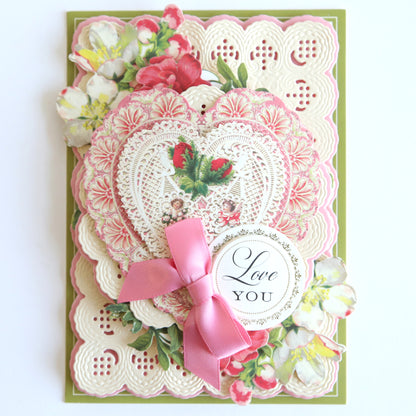 A decorative card featuring layered lace patterns and vintage botanicals, embellished with a pink ribbon and a tag reading "Love You," exudes charm. Intricate lace doilies enhance the design, making it an elegant piece perfect for enthusiasts of Love Ephemera Stickers.