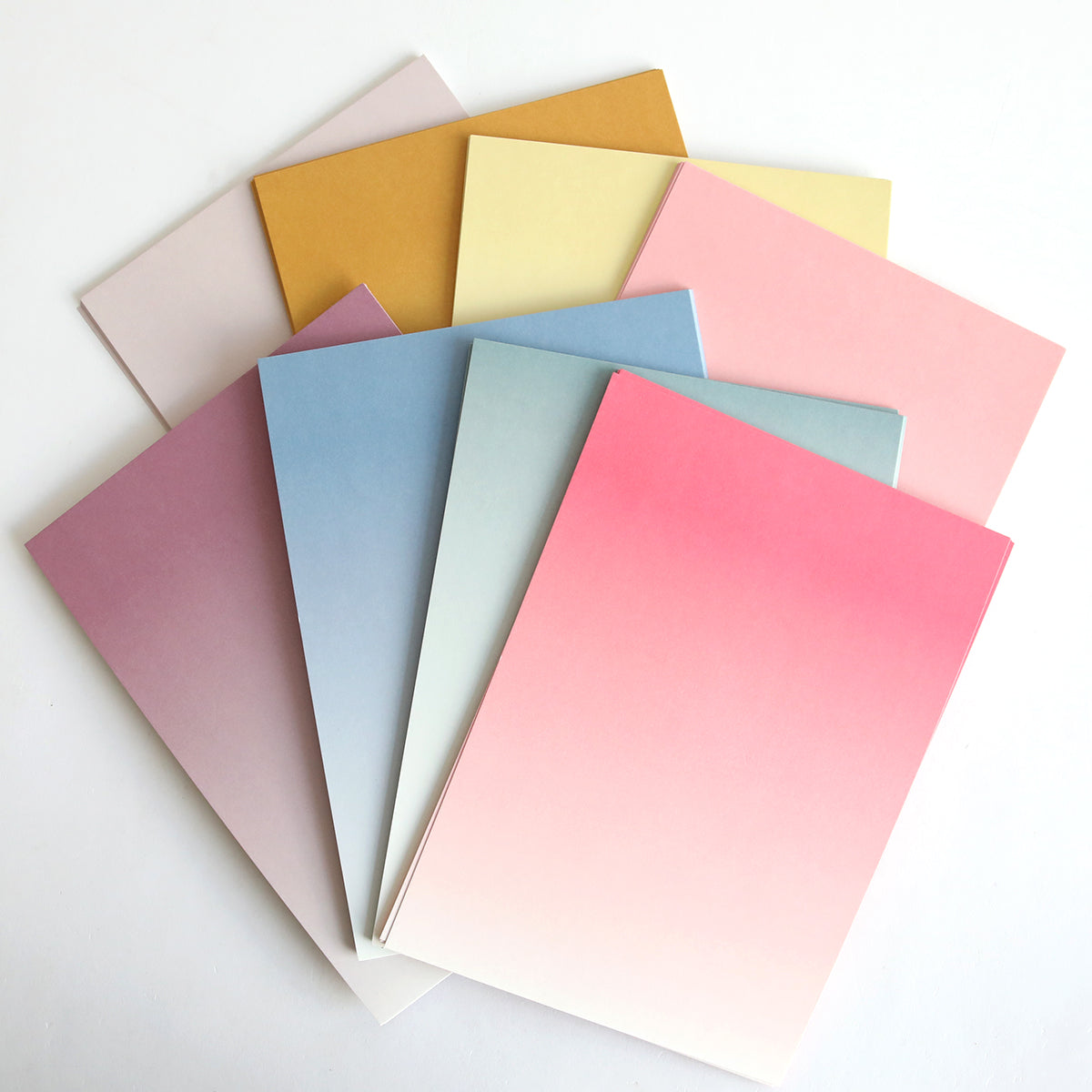 Seven Ombre Double Sided Cardstock sheets, featuring gradient shades of pink, blue, yellow, and beige, are fanned out on a white surface—ideal for adding a touch of elegance to paper crafts.