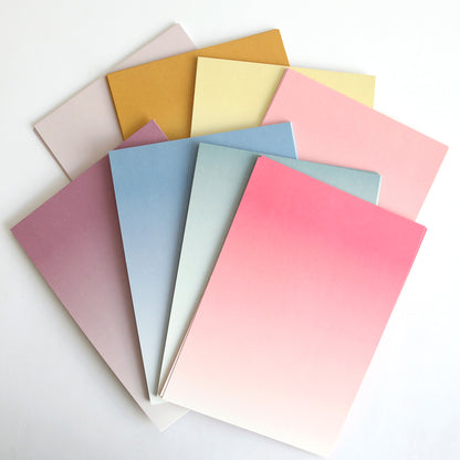 Seven Ombre Double Sided Cardstock sheets, featuring gradient shades of pink, blue, yellow, and beige, are fanned out on a white surface—ideal for adding a touch of elegance to paper crafts.