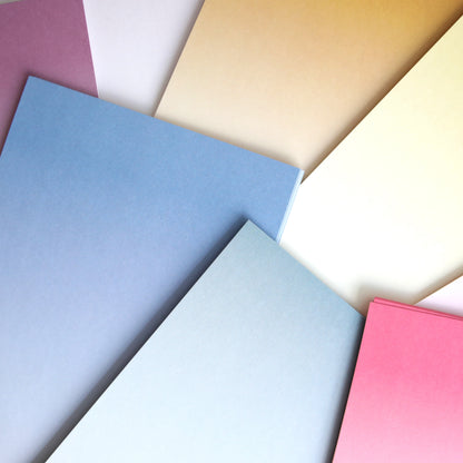 Ombre Double Sided Cardstock sheets in blue, pink, yellow, and white are fanned out on a surface—ideal for paper crafts.