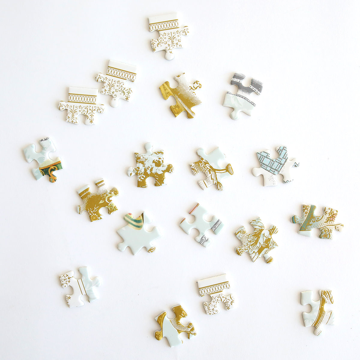 A delightful mix for tea and puzzle lovers, the High Tea Jigsaw Puzzle's scattered white and gold pieces lay on a white surface.