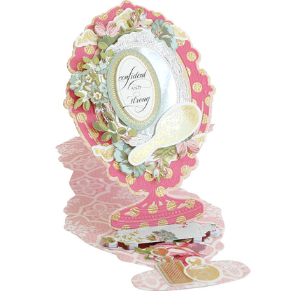 The Mirror Card Craft Box includes an intricate pop-up card made with double-sided cardstock. It showcases a decorative mirror design adorned with pink and floral patterns and foil embellishment stickers. The phrase "confident and strong" adds empowerment to the elegant card.