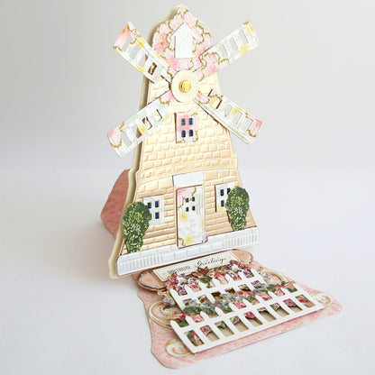 The Windmill 12x12 Cardstock is a pop-up greeting card featuring a 3D windmill with white blades and a pink wall, crafted from double-sided cardstock, and accented by bushes and a decorative fence.