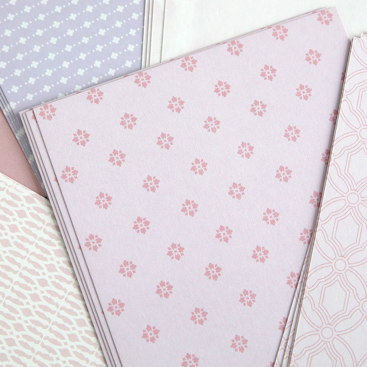 Stacks of pastel-patterned paper with pink floral and geometric designs overlap, perfect for card making. Ideal for use with Color Drenched Cardstock Fresh Lilac and hot foil plates.