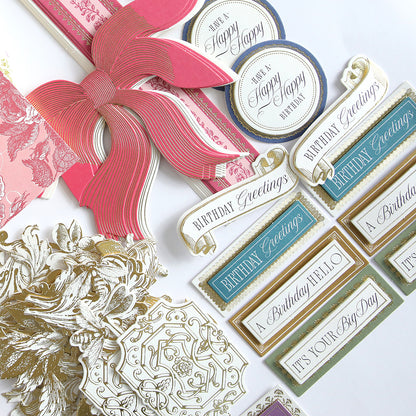 A beautifully arranged Birthday Present Card Making Kit is displayed on a white surface, featuring assorted handmade birthday cards, tags, and embellishments in gold, pink, blue, and white. The set includes a large paper bow and intricate floral designs for decorating your creations.