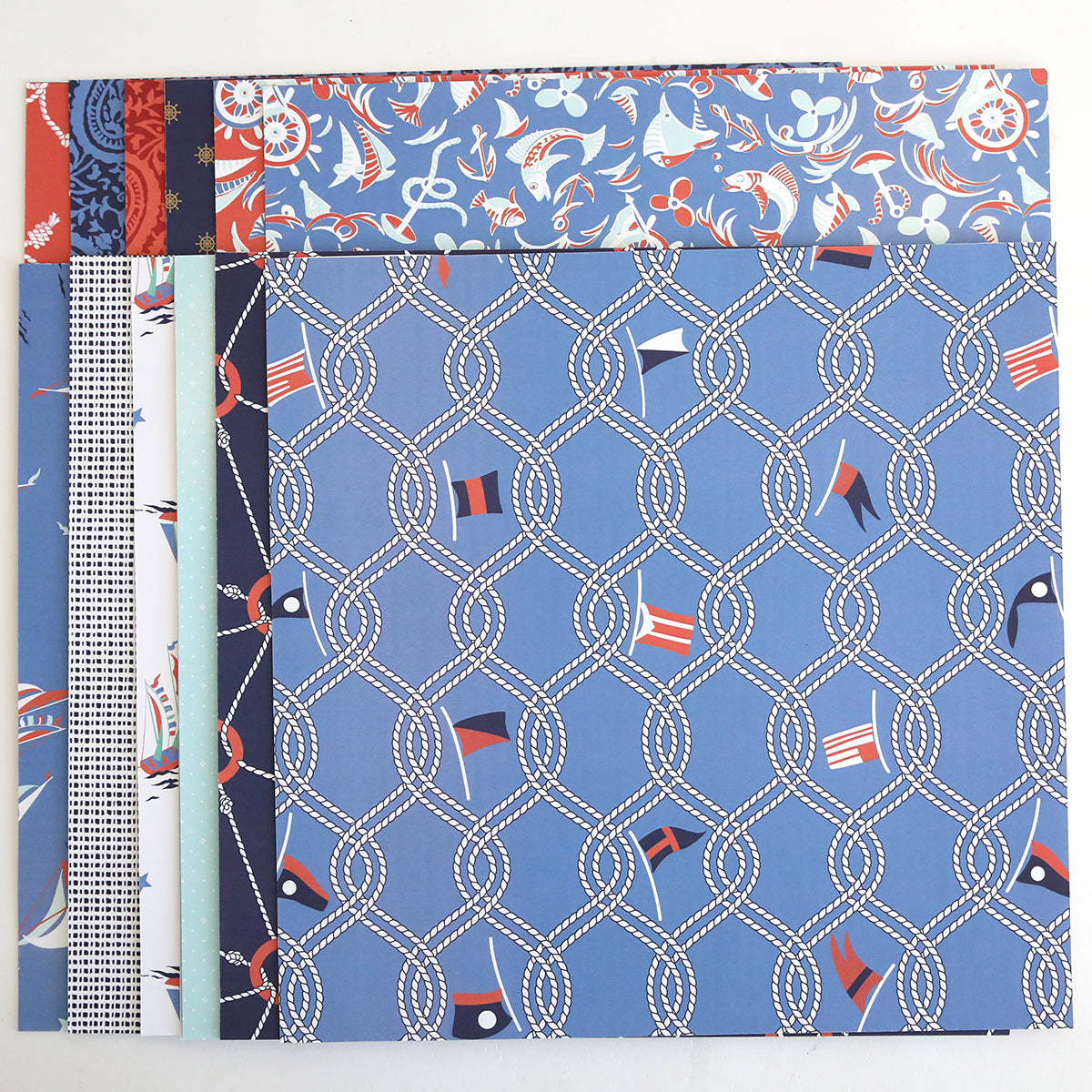 The Lighthouse 12x12 Cardstock features nautical patterns—ropes, flags, boats, and anchors—displayed in overlapping layers on a white background. Ideal for pairing with Lighthouse Easel Dies in your next craft project.