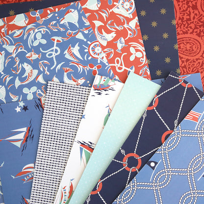 Lighthouse 12x12 Cardstock features double-sided sheets with nautical patterns—ships, anchors, ropes, stars, and geometric prints—in blue, navy, red, and white. Ideal for scrapbooking or pairing with Lighthouse Easel Dies.