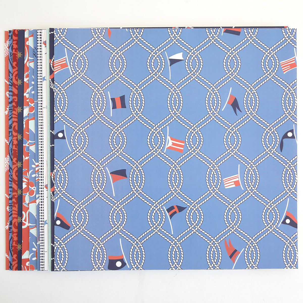 Lighthouse 12x12 Cardstock: Stack of double-sided sheets with coastal designs, featuring nautical motifs like ropes and signal flags in blue, red, and white, arranged side by side on a white background.
