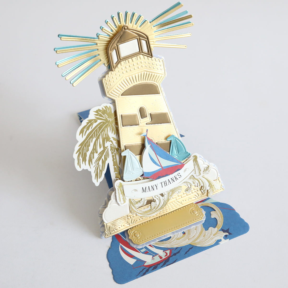 A pop-up greeting card showcases a gold lighthouse, sailboats, palm tree, and "Many Thanks" on a blue and white coastal design base made with premium Lighthouse 12x12 Cardstock.