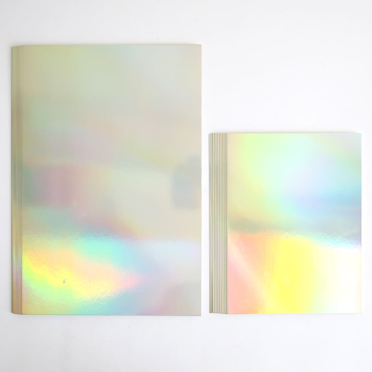 Two Luxury Iridescent Foil Cardstock notebooks rest on a white surface; the larger one is on the left, while smaller stacked notebooks display a brilliant silver holographic foil finish.