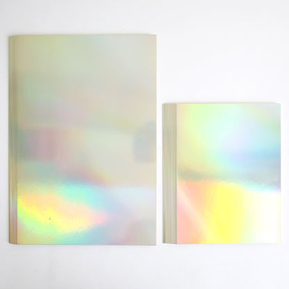 Two Luxury Iridescent Foil Cardstock notebooks rest on a white surface; the larger one is on the left, while smaller stacked notebooks display a brilliant silver holographic foil finish.