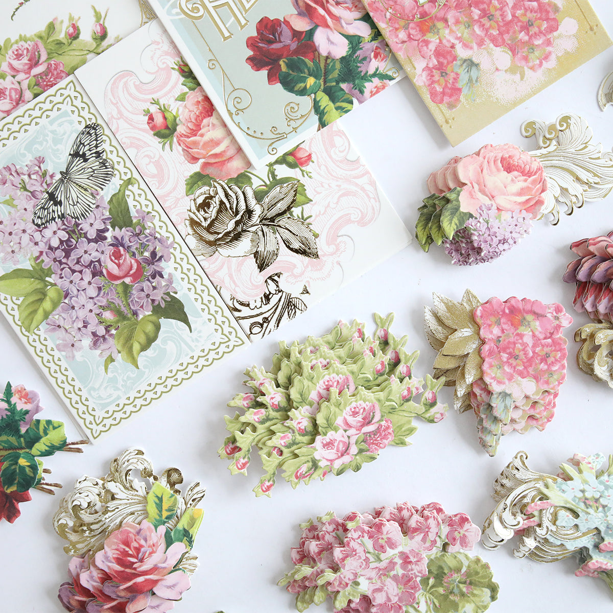 Antique Greetings Postcard Embellishment Stickers featuring roses, hydrangeas, butterflies, and ornate gold accents are artfully displayed on a white surface.