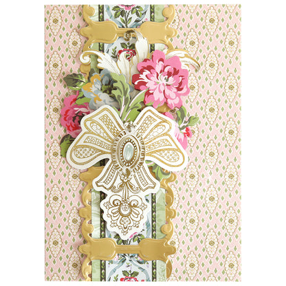 A decorative greeting card crafted with 12″ Regal Trimmings Border Dies and rose gold matte foil cardstock, featuring a large gold bow, pink and green floral accents, and an ornate patterned background.