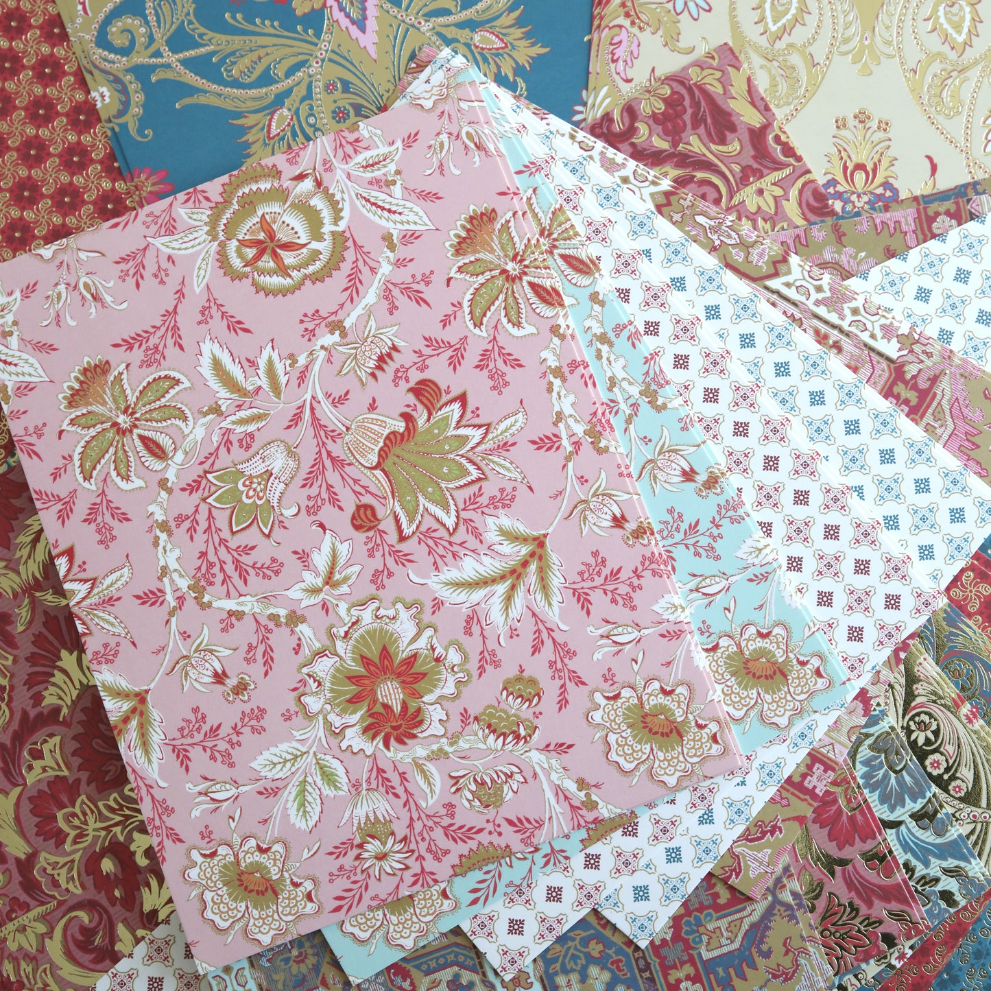 Several sheets from the Jasmine Cardstock Collection, featuring intricate floral and geometric designs in pink, blue, and shimmering gold foil cardstock, are layered together.