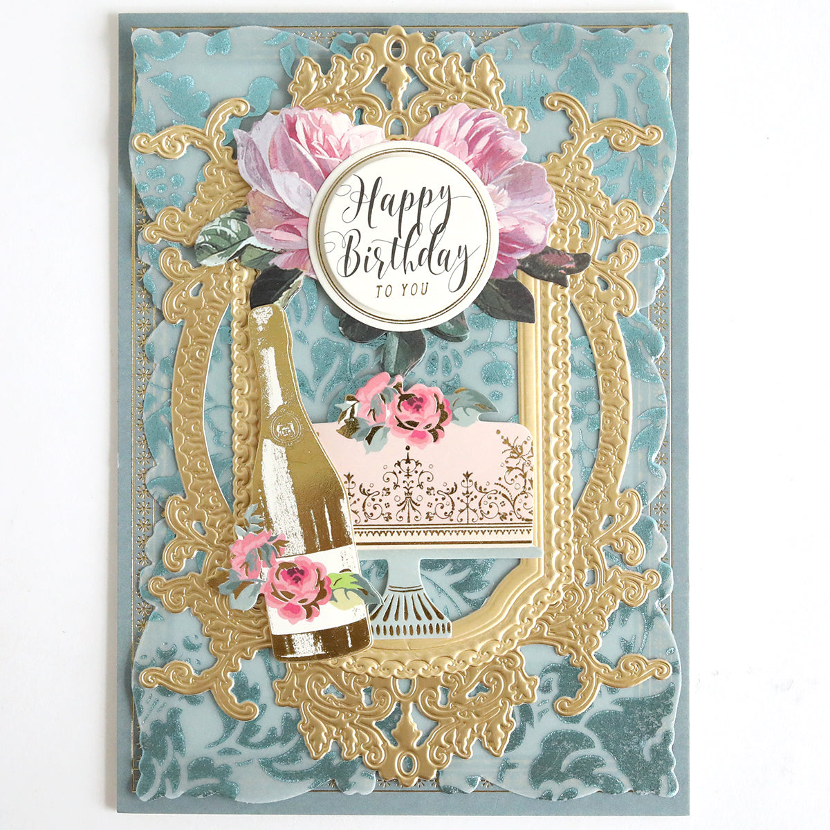 The “Rose and Pineapple Gilded Hot Foil Plates with Perfect Palette Pastel Hot Foil” creates a decorative birthday card featuring gold ornate frames, a pink cake on a stand, champagne bottle, pink flowers, and a “Happy Birthday to You” message set against a floral background—an elegant combination of gilded crafts using pastel foil accents.