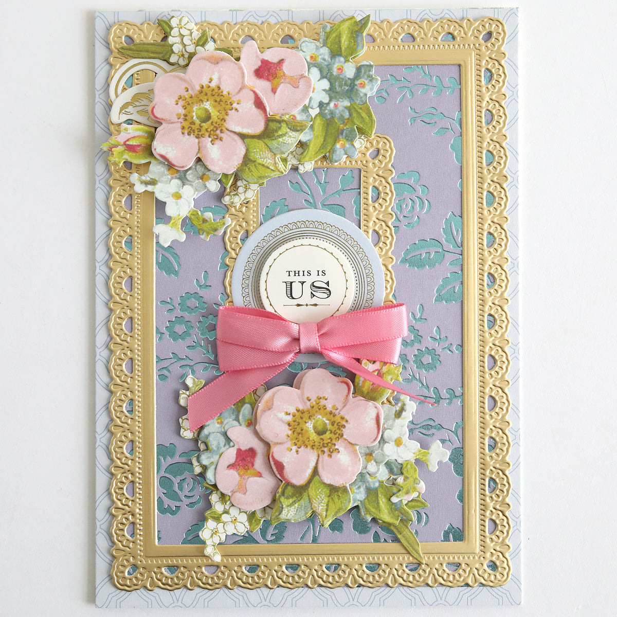 A decorative greeting card features designs created with the Rose and Pineapple Gilded Hot Foil Plates accented by Perfect Palette Pastel Hot Foil, adorned with pink floral motifs, gold foil highlights, a pink ribbon embellishment, and a central “This is Us” emblem set against a lavender patterned background.