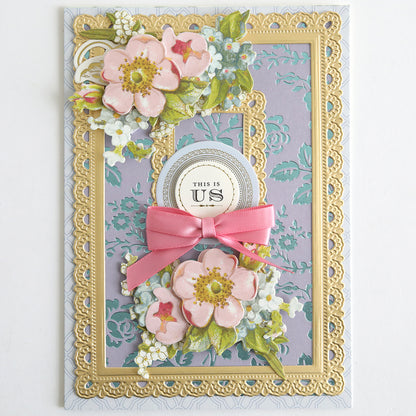 A decorative greeting card features designs created with the Rose and Pineapple Gilded Hot Foil Plates accented by Perfect Palette Pastel Hot Foil, adorned with pink floral motifs, gold foil highlights, a pink ribbon embellishment, and a central “This is Us” emblem set against a lavender patterned background.