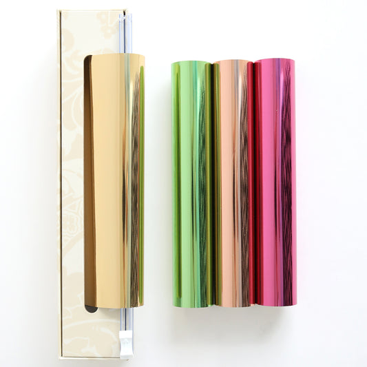Four rolls of Perfect Palette Antique Gild Hot Foil—gold, green, copper, and pink—are shown by a patterned box on a white surface, ideal for adding shimmering accents to your paper crafts.