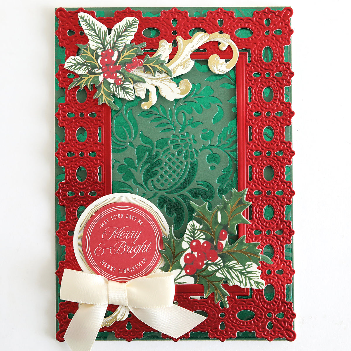 A festive card made using the Empress Gild Hot Foil Machine with Holiday Hot Foil, featuring a red lace-inspired border, holly and berry motifs, gold foiled accents, a “Merry & Bright” greeting, and topped with a cream ribbon bow.