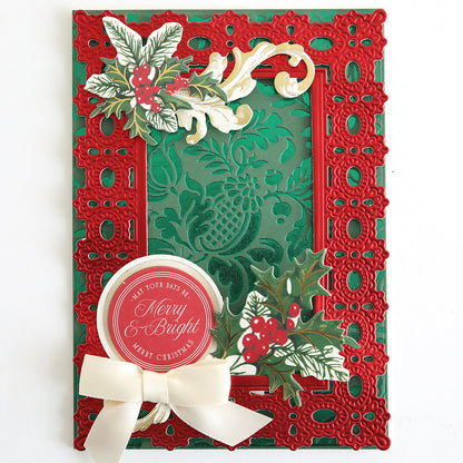 A festive card made using the Empress Gild Hot Foil Machine with Holiday Hot Foil, featuring a red lace-inspired border, holly and berry motifs, gold foiled accents, a “Merry & Bright” greeting, and topped with a cream ribbon bow.