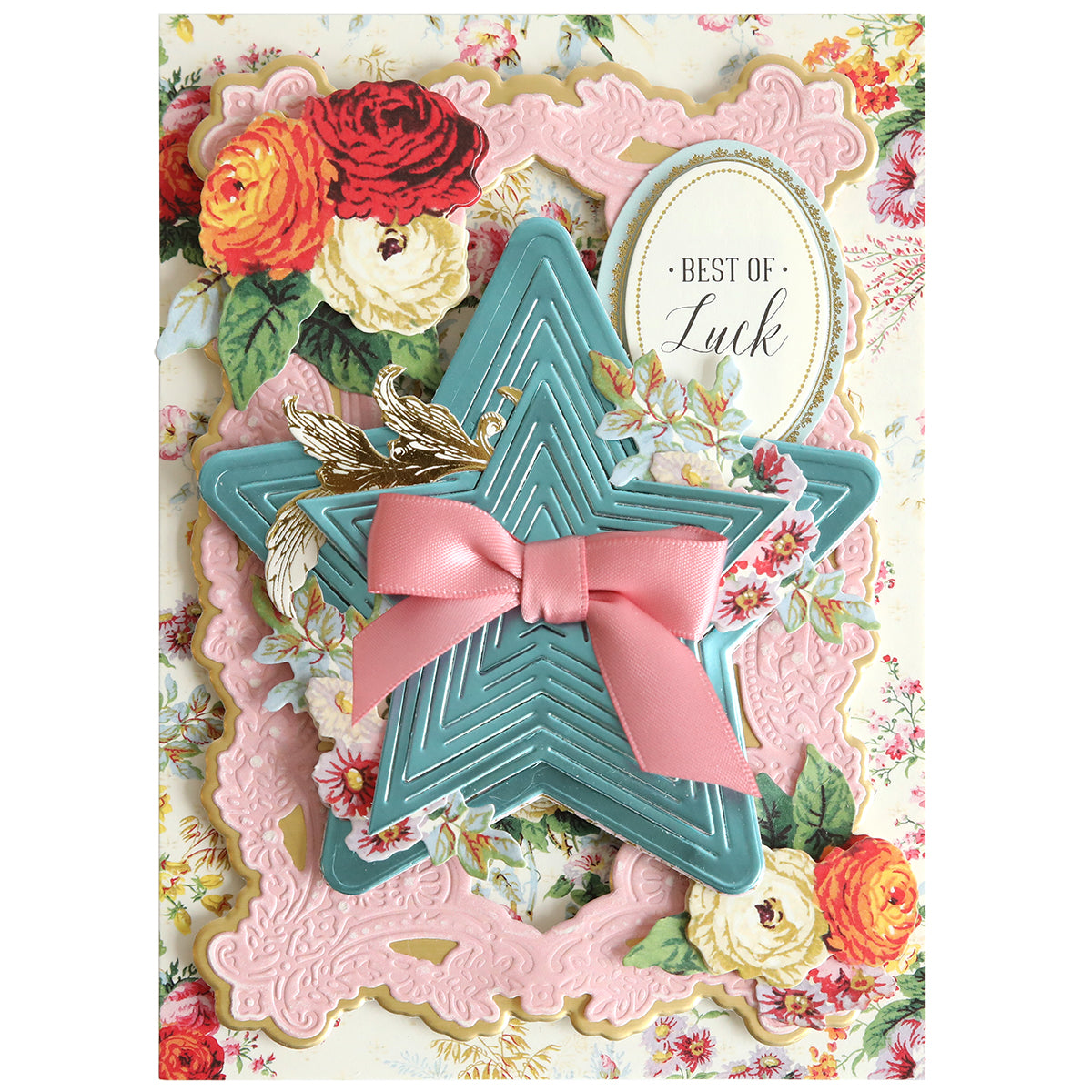 A decorative card featuring layered floral patterns, a pink bow on dimensional stars cut with the Kirigami Star Dies, gold leaves, and an oval label that says “Best of Luck.”.