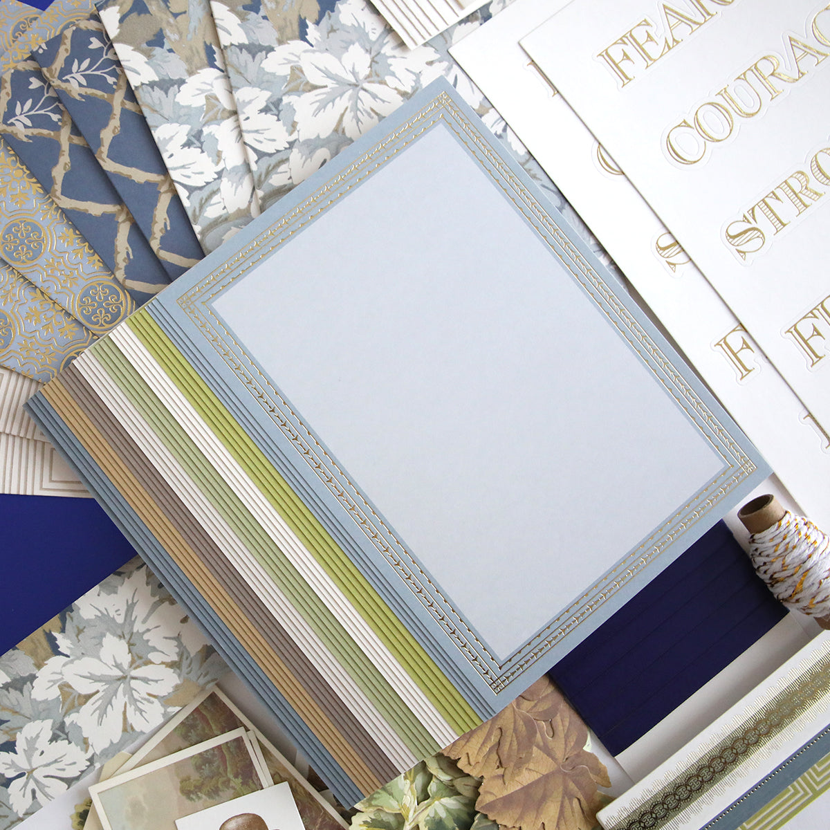 A flat lay featuring Anna Griffin | Create Spring 2026 patterned papers, a striped-spine notebook, gold-printed word sheets, and blue, green, and neutral accents—ideal inspiration for your next scrapbooking event.