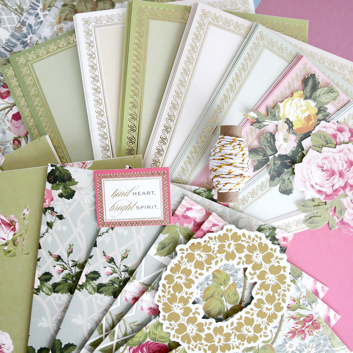 The Rose Ultimate Paper Crafting Collection features assorted floral-patterned stationery and envelopes in pink, green, and gold—ideal for card making and paper crafting—with decorative frames, twine, and a "kind heart. bright spirit." card.