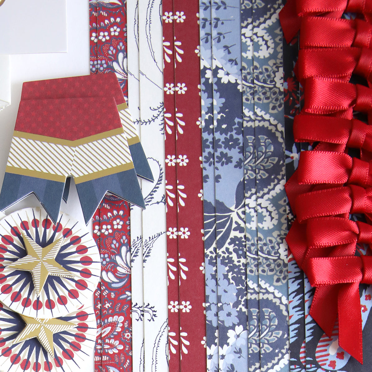 Patterned blue and red paper strips, red ribbons, and decorative cut-outs from the Patriotic Papercrafting Kit with Dies featuring geometric and floral designs are arranged neatly on a white surface.