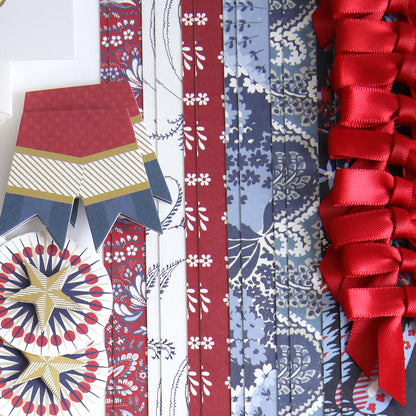 Patterned blue and red paper strips, red ribbons, and decorative cut-outs from the Patriotic Papercrafting Kit with Dies featuring geometric and floral designs are arranged neatly on a white surface.