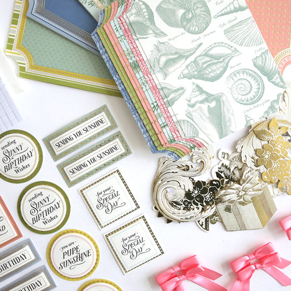 The Simply Summer Card Making Kit displays decorative paper, labels, die-cut shapes, and pink ribbon bows arranged on a white surface for crafting handmade cards.