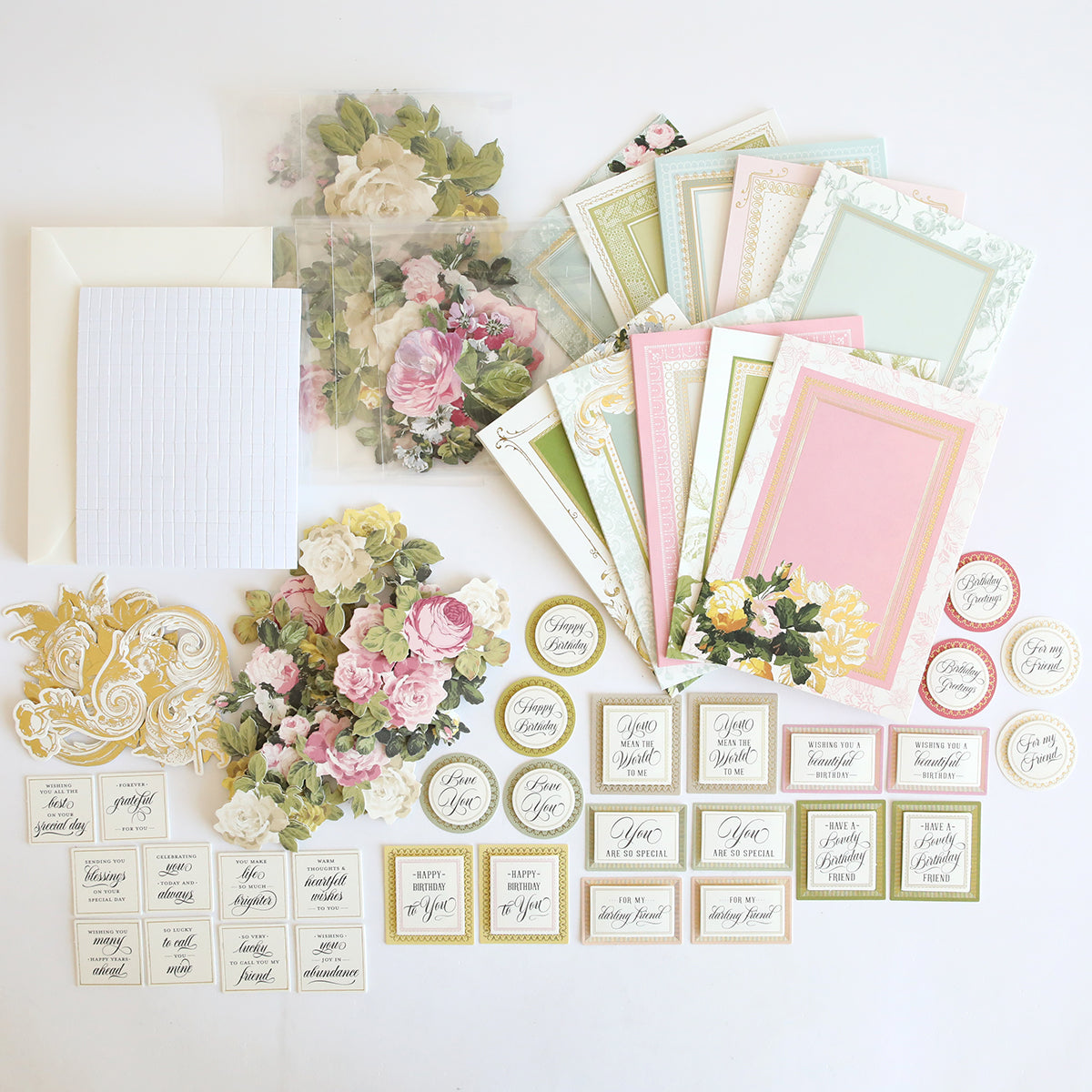 The Folded Rose Card Making Kit features pastel greeting cards, 3D embellishments, floral die-cuts, envelopes, and assorted sentiment labels arranged on a white surface.