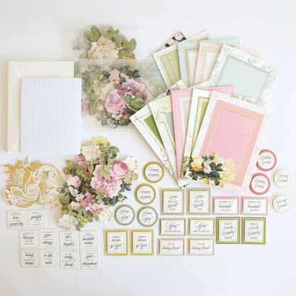 The Folded Rose Card Making Kit features pastel greeting cards, 3D embellishments, floral die-cuts, envelopes, and assorted sentiment labels arranged on a white surface.