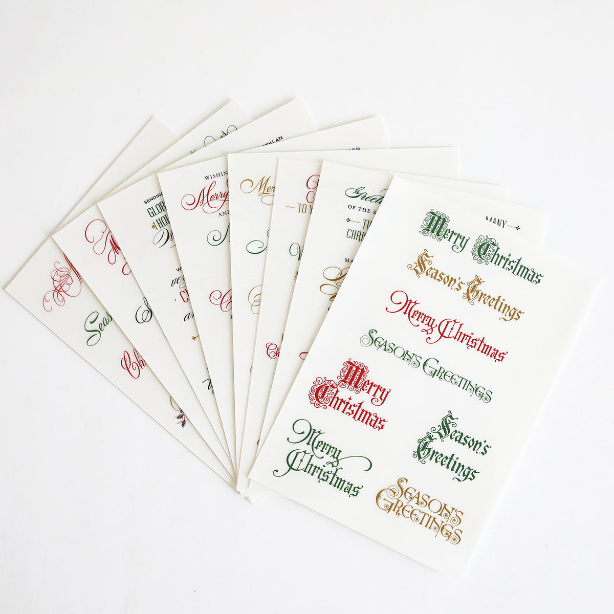 Eight sheets of the Christmas Card Sentiment Rub Ons are fanned out on a white background, each featuring festive "Merry Christmas" and "Season's Greetings" phrases in assorted fonts and colors—ideal for holiday paper crafts or enhancing handmade cards with a cheerful touch.