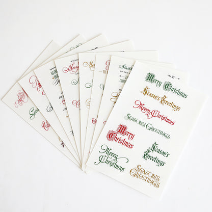 Eight sheets of the Christmas Card Sentiment Rub Ons are fanned out on a white background, each featuring festive "Merry Christmas" and "Season's Greetings" phrases in assorted fonts and colors—ideal for holiday paper crafts or enhancing handmade cards with a cheerful touch.