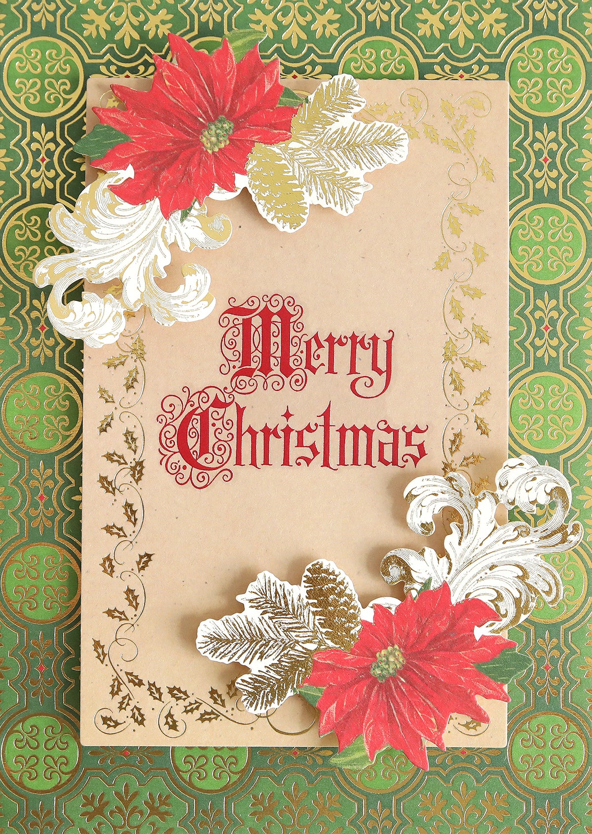 A Christmas card adorned with red poinsettias, gold leaves, and "Merry Christmas" in red on a brown background sits on a green patterned surface—a wonderful handmade creation to elevate your holiday crafts. Complete your design with Christmas Card Sentiment Rub Ons for an extra festive touch.