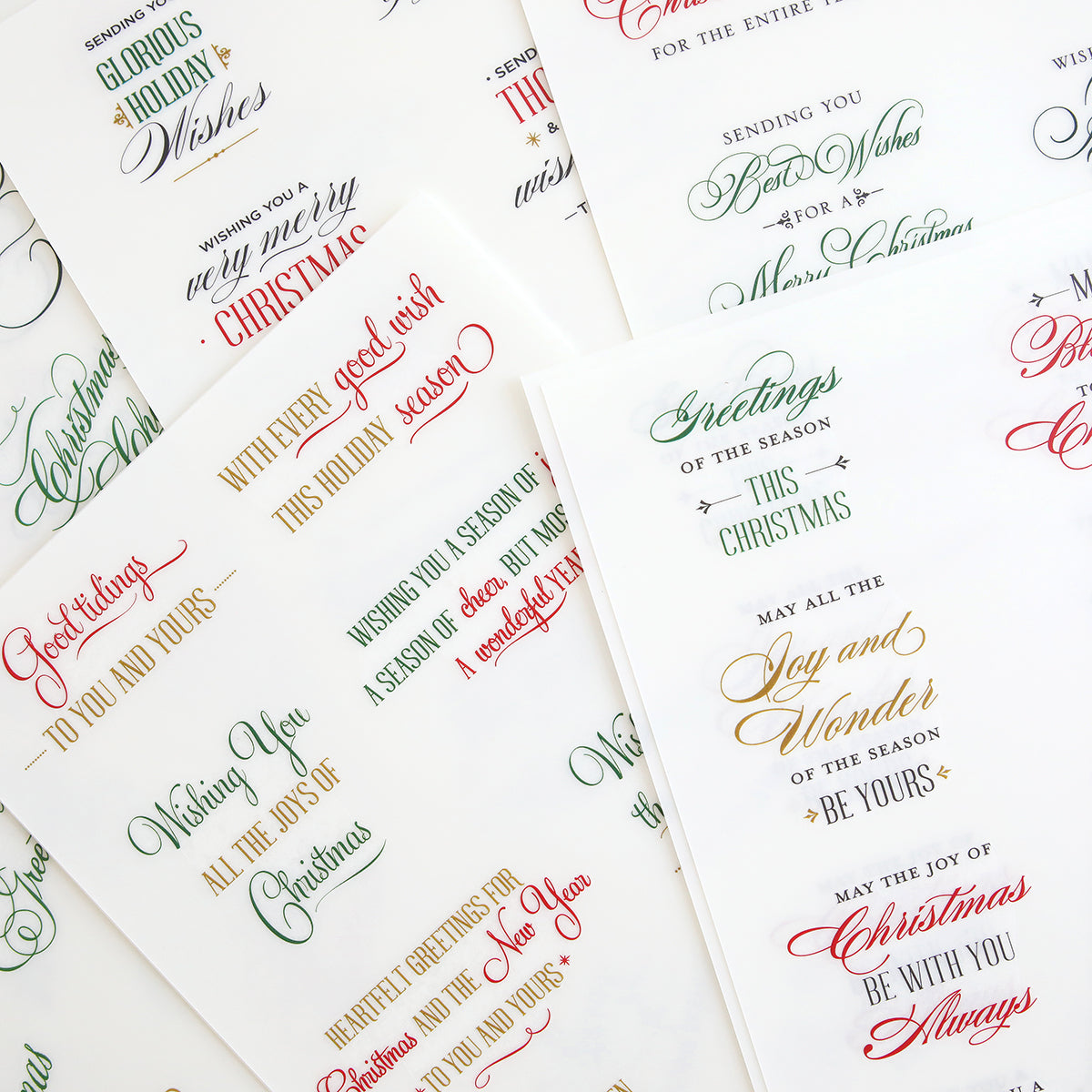 Christmas Card Sentiment Rub Ons—assorted sheets featuring festive Christmas greetings in various fonts and colors like red, green, gold, and black, overlapping on a white background; ideal for embellishing handmade cards or adding to holiday paper crafts.