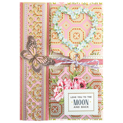 Greeting card featuring ornate patterns, a floral heart, butterfly, and flowers, tied with a ribbon. Expertly crafted using layered paper techniques and the Charlie Double Sided 12x12 Cardstock Collection for added dimension. A label displays “LOVE YOU TO THE MOON AND BACK” in striking black and gold text.